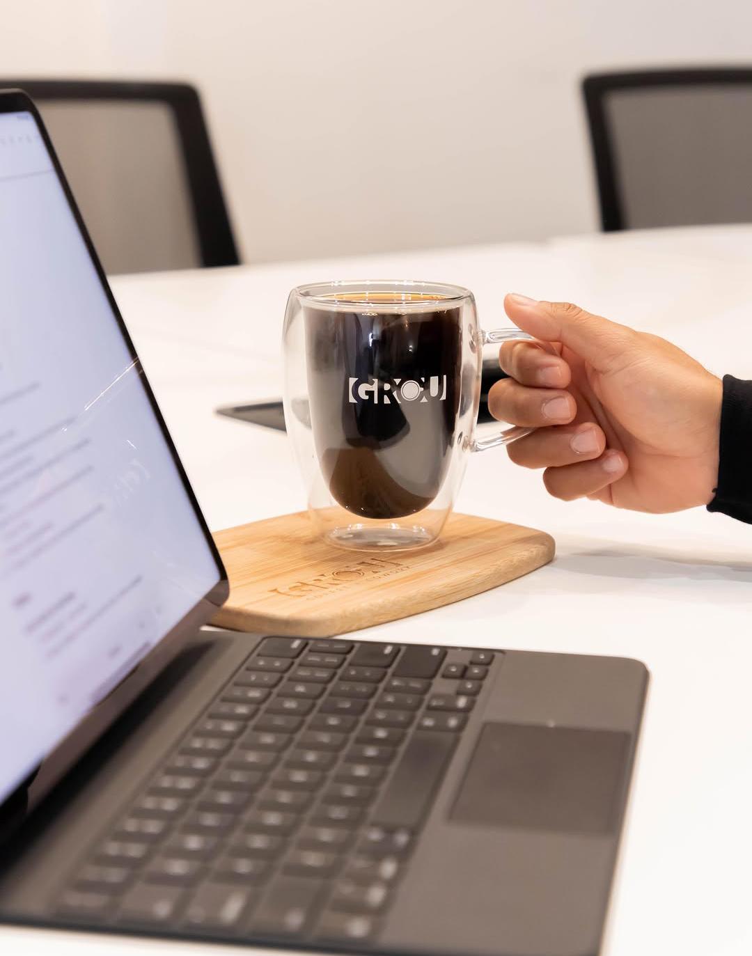 Hand holding the handle of a GROU glass mug of black coffee on a wooden board beside a laptop