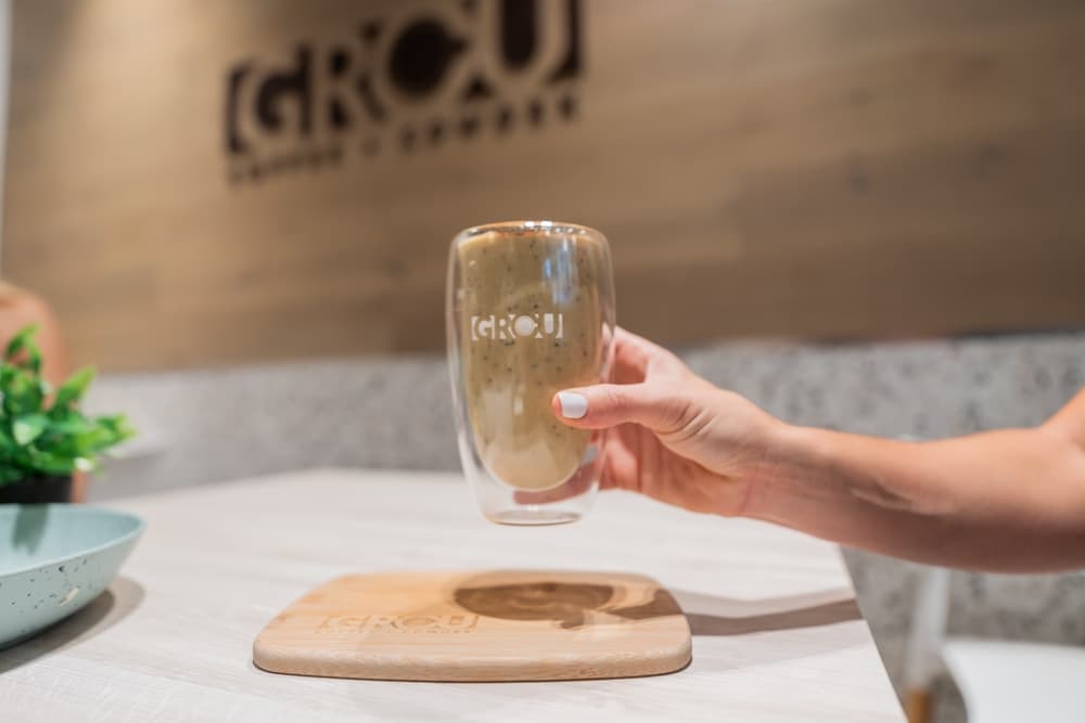 Mano sosteniendo el Hormone Harmony Smoothie en un vaso de GROU sobre una tabla de madera, con el logo de GROU en la pared