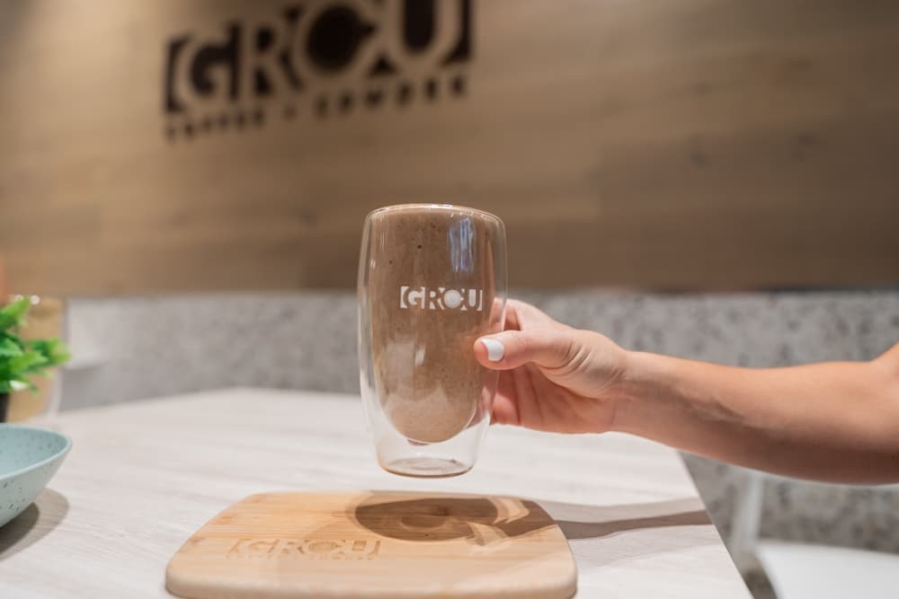Mano sosteniendo el Muscle Repair smoothie, espeso y marrón, en un vaso de GROU sobre una tabla de madera, con el logo de GROU detrás