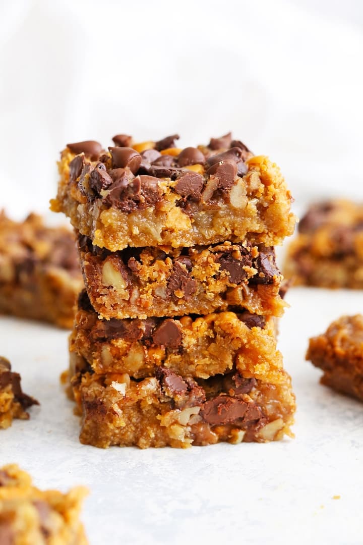 Stack of four Dolly bars with chocolate chips and chopped nuts, more bars scattered around on a white surface