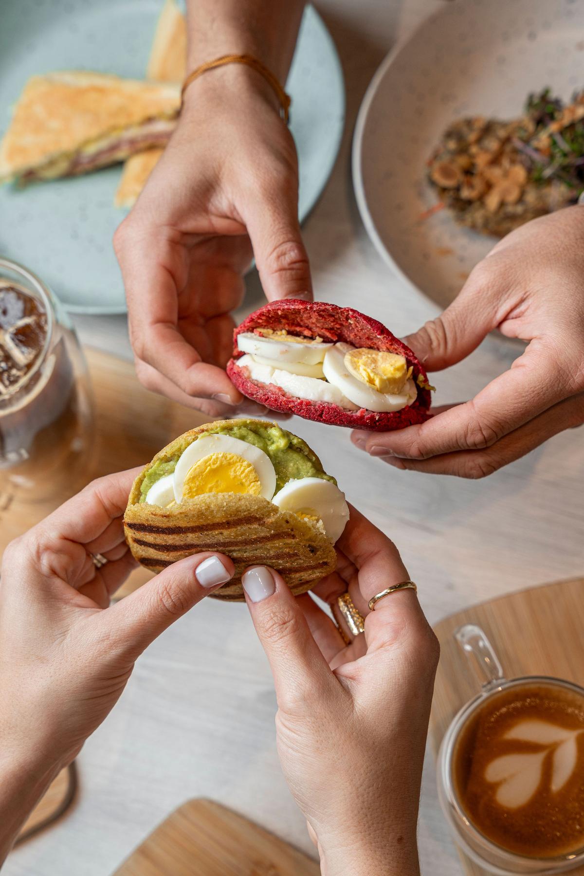 Hands holding a beet arepa with boiled egg and a grilled plantain arepa with avocado and egg at GROU, beside a latte