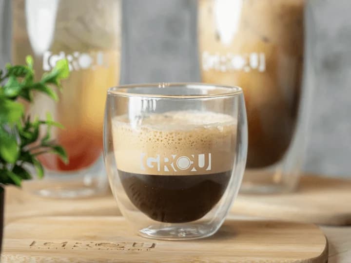 Vaso pequeño de GROU con café y una capa gruesa de espuma sobre una tabla de madera, con dos bebidas en vasos altos de GROU desenfocadas al fondo