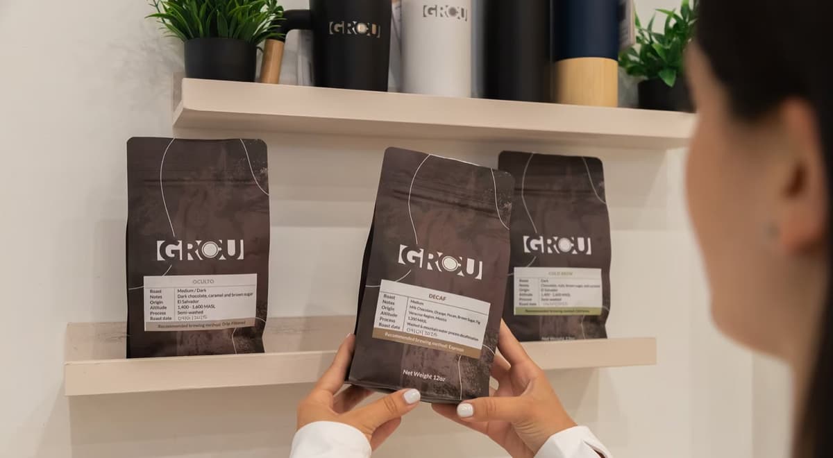 Hands holding a GROU Decaf coffee bag in front of GROU Oculto and Cold Brew bags on a shelf