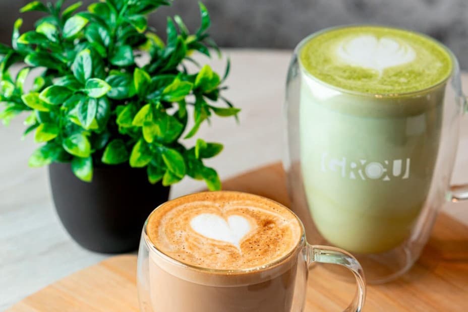 Latte con arte en forma de corazón junto a un matcha latte en taza de vidrio GROU y una pequeña planta en maceta