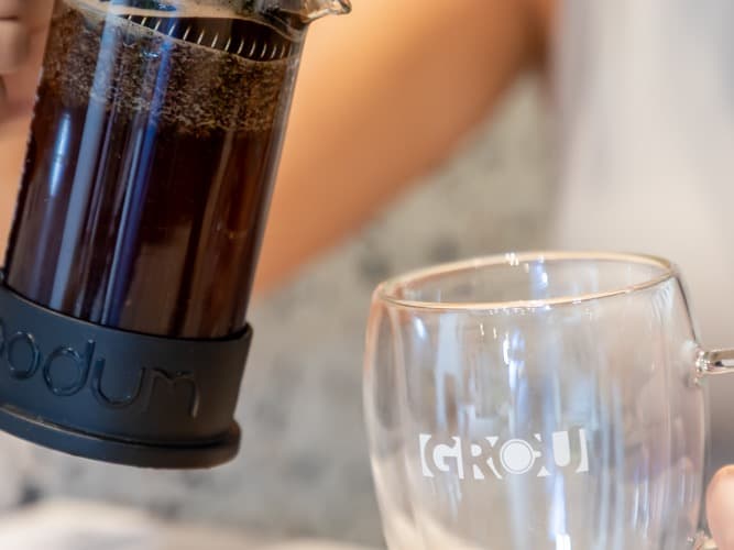 French press of brewed coffee tilted to pour into a GROU glass mug