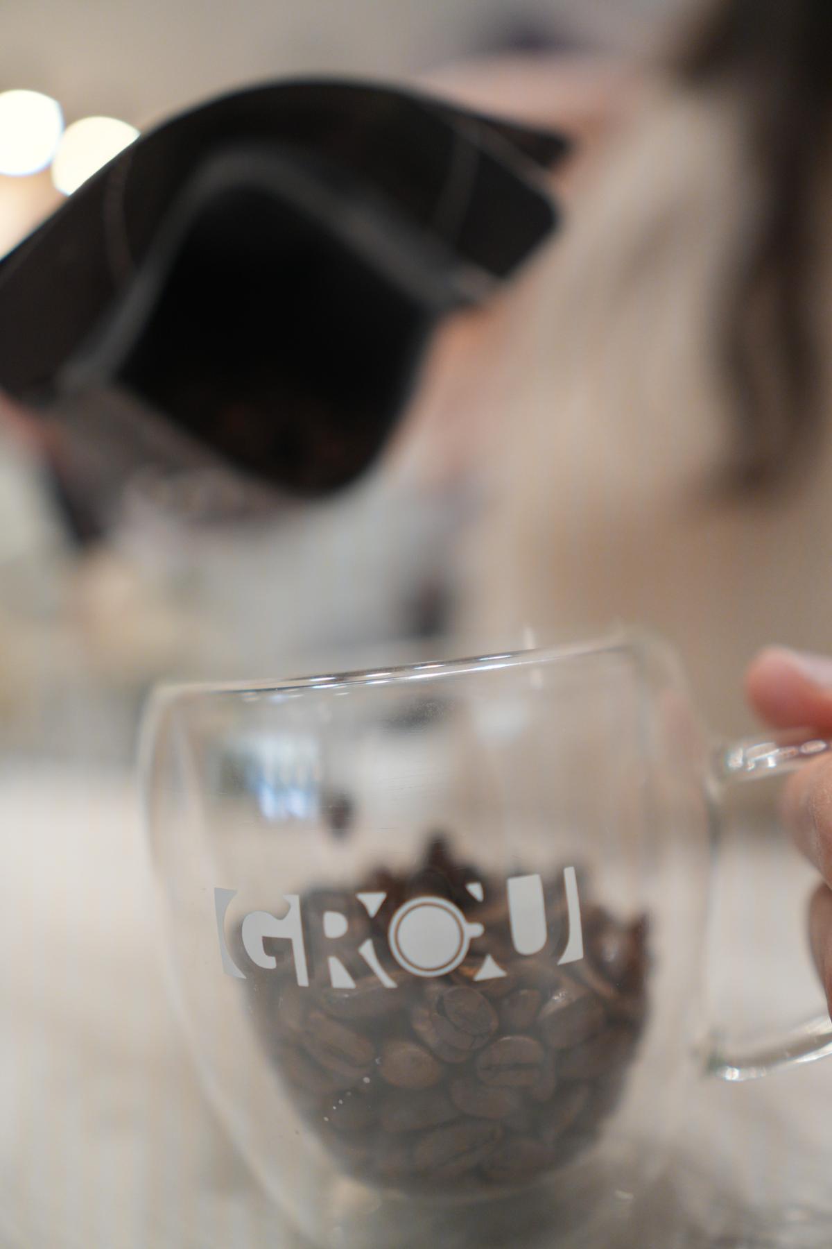 Roasted coffee beans poured from a dark bag into a clear glass mug with the GROU logo