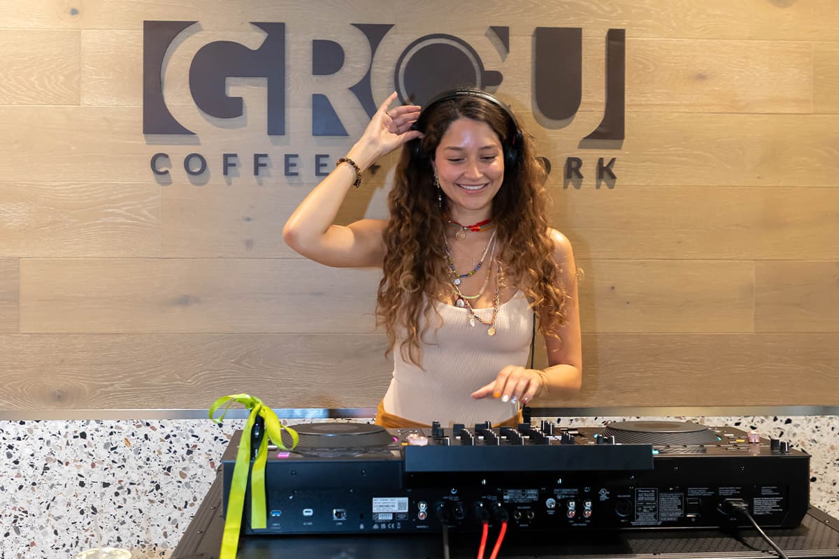 Smiling DJ in headphones at a DJ controller in front of the GROU Coffee + Cowork wall sign