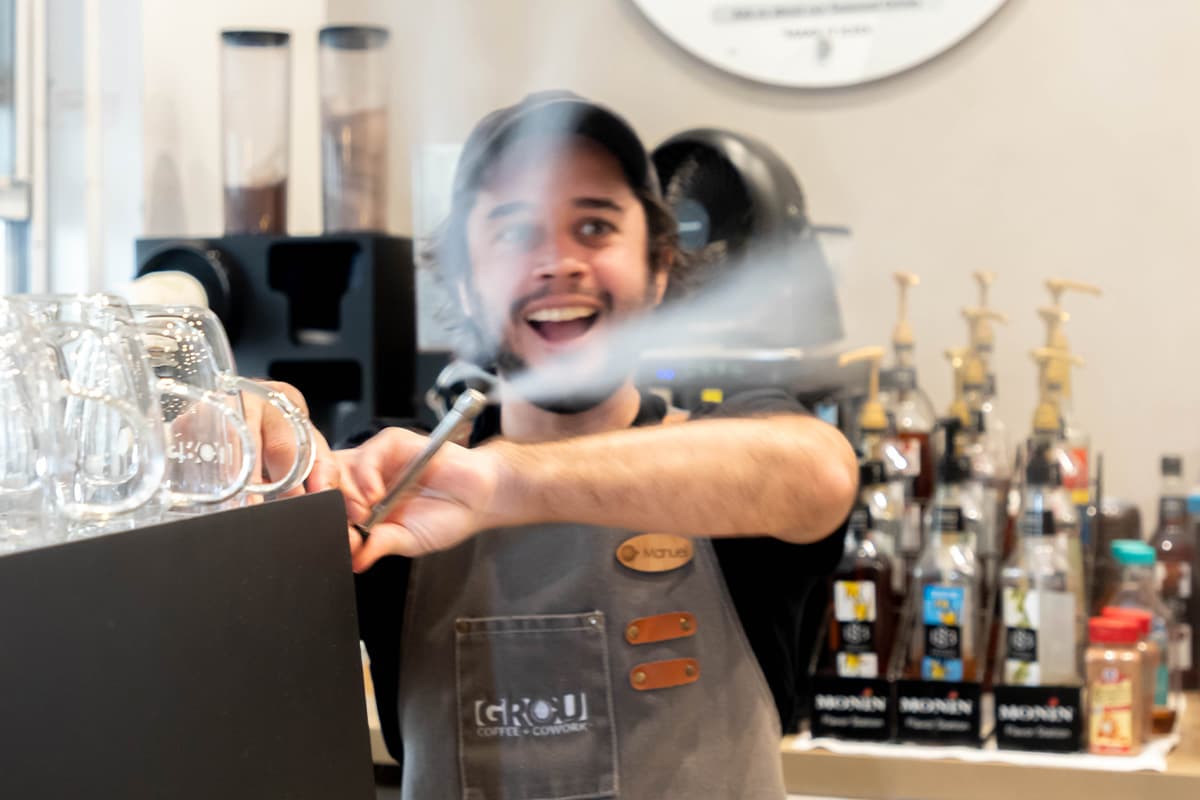Smiling GROU barista in a branded apron purging the espresso machine steam wand behind the bar
