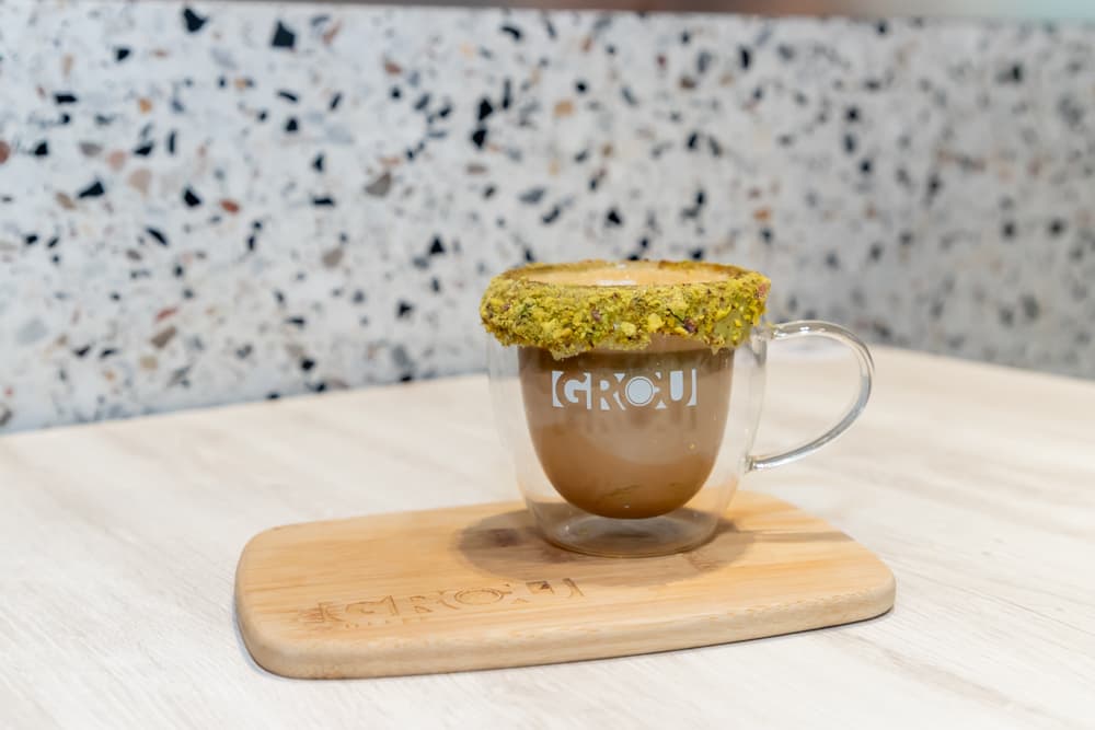 Pistachio Cappuccino con borde de pistachos triturados en una taza de vidrio de GROU sobre una tabla de madera