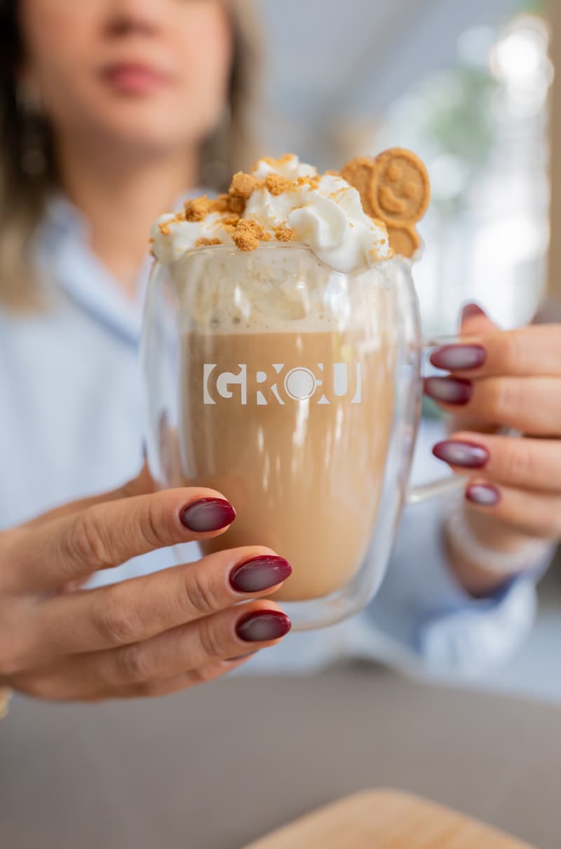 Manos sosteniendo un Gingerbread Latte en una taza de vidrio GROU con crema batida, migas doradas y una galleta con carita feliz