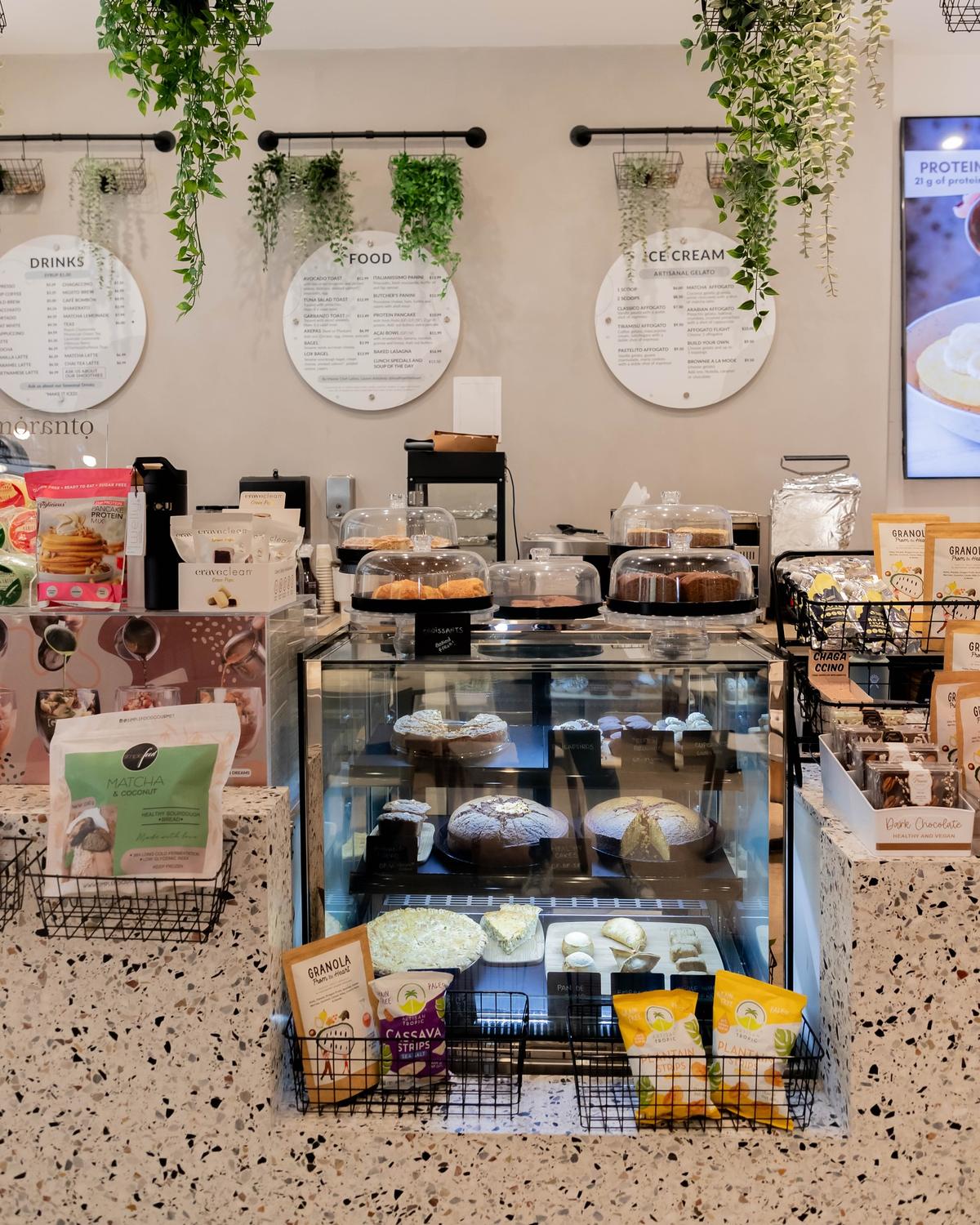 Mostrador de GROU con los menús de bebidas, comida y helados, plantas colgantes, una vitrina de repostería y snacks empacados