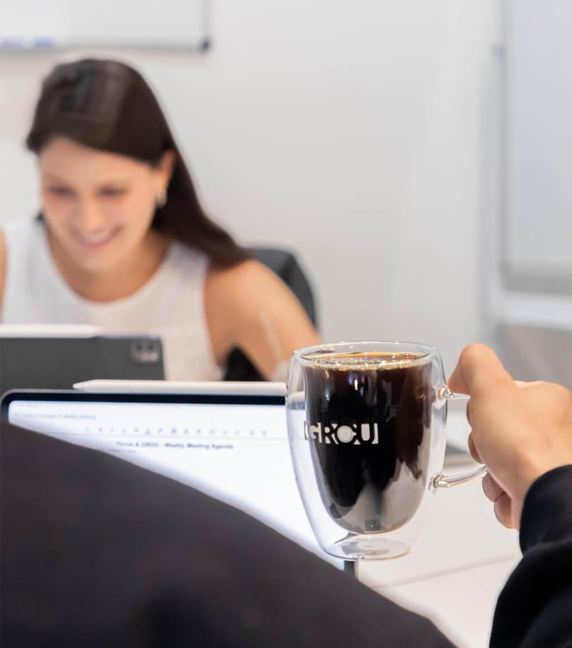 Mano sosteniendo una taza de vidrio GROU con café negro mientras una mujer sonríe frente a su pantalla trabajando en GROU