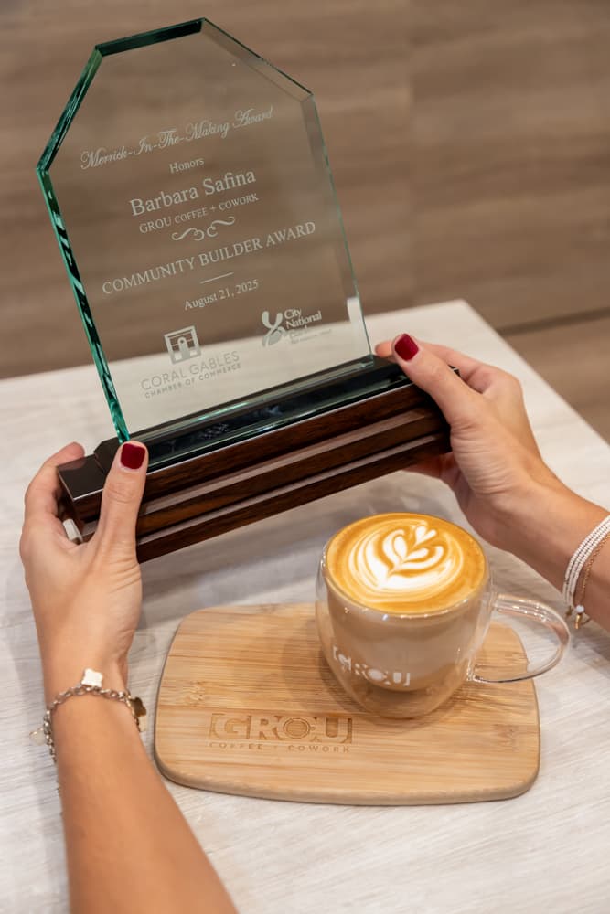 Trofeo de cristal del Community Builder Award para Barbara Safina, sostenido junto a un café con latte art sobre una tabla de madera GROU