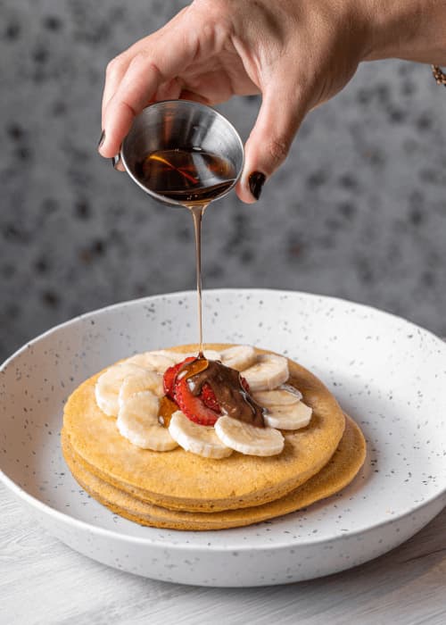 Pila de protein pancakes de GROU con banana y fresas en rodajas mientras se vierte sirope desde una tacita