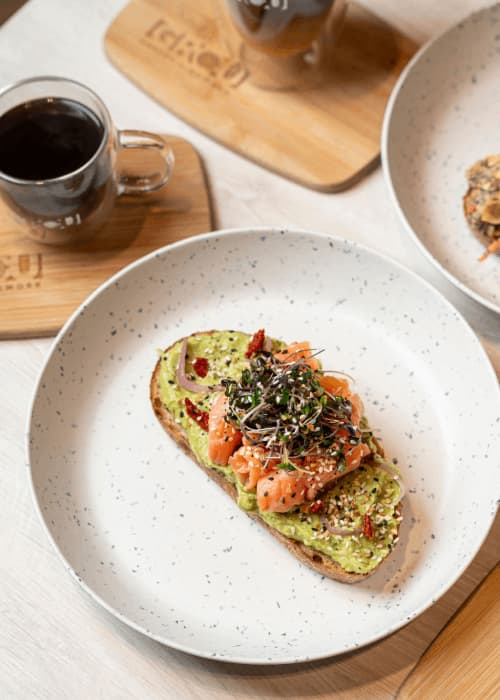 Avocado toast en pan multigrano con salmón, brotes y semillas, junto a un café negro en GROU