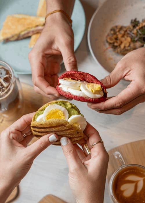 Manos sosteniendo una arepa de remolacha con huevo duro y una de plátano con aguacate y huevo, junto a un latte
