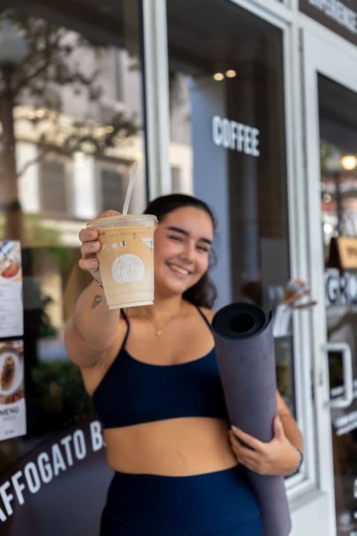 Mujer con ropa deportiva mostrando una bebida fría en un vaso de GROU y una colchoneta enrollada frente al coffee shop