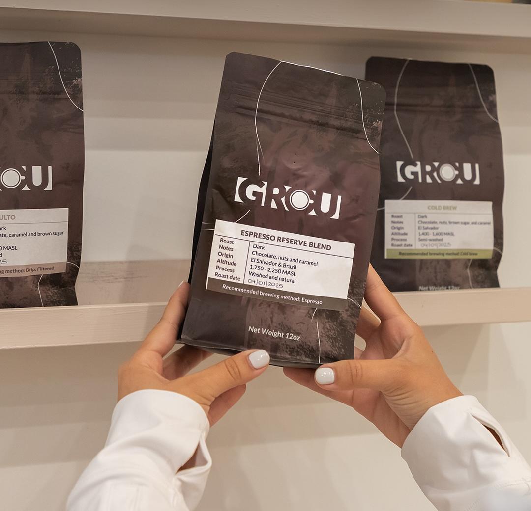 Hands holding a bag of GROU Espresso Reserve Blend coffee in front of a shelf with more GROU coffee bags